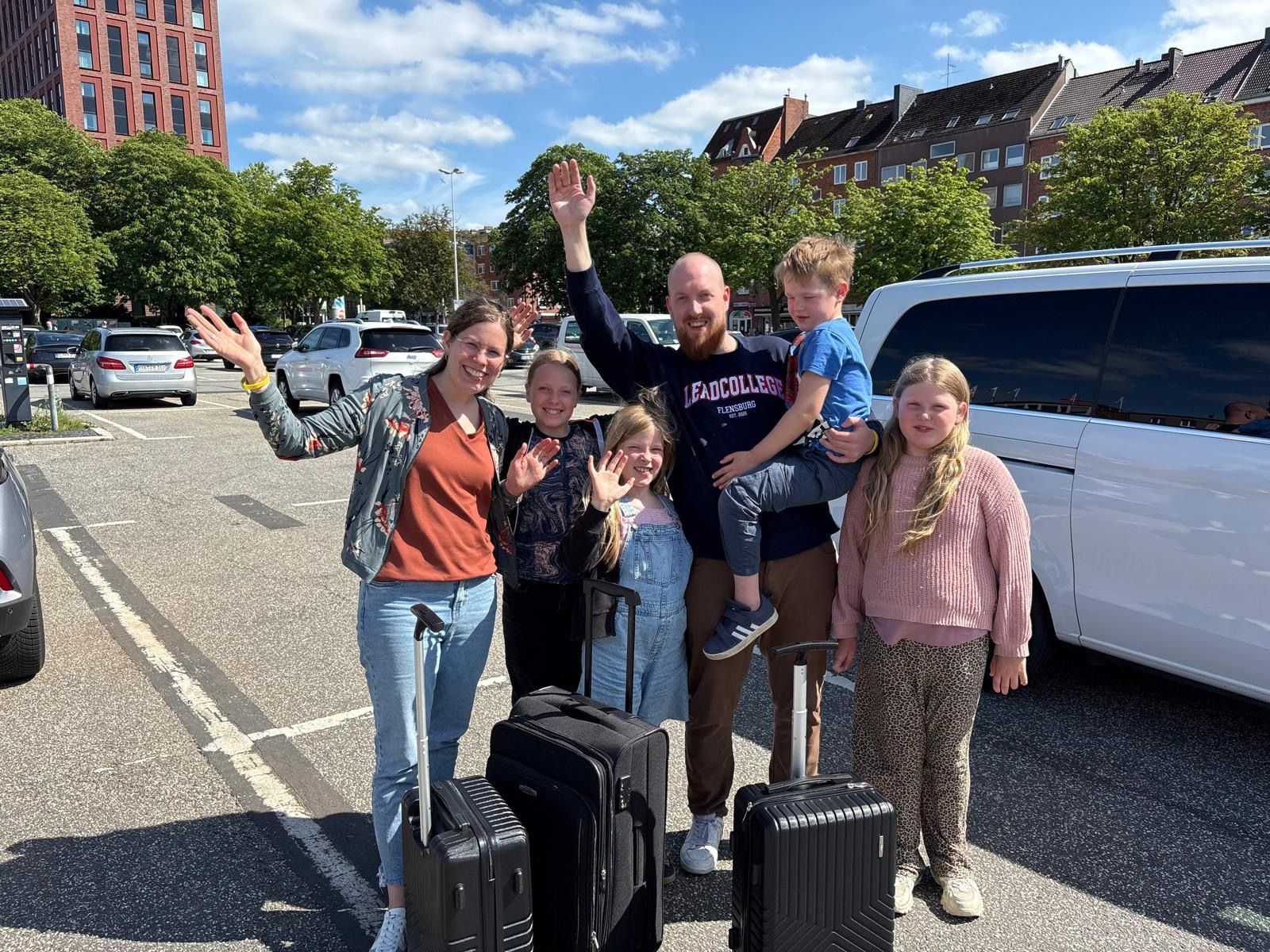 Christoph und Rebecca mit Antonia, Louisa, Paulina und Jakob – mit Koffern, lachend und winkend auf einem sonnigen Platz in Kiel.